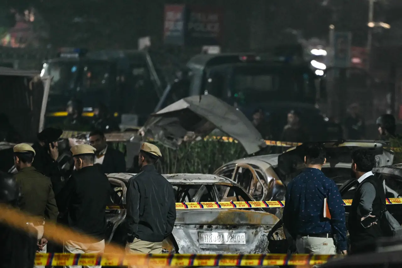 Police and security personnel stand behind the safety cordons delimiting the blast site as they inspect charred vehicles following an explosion near the Red Fort, in the old quarters of Delhi, on November 10, 2025.